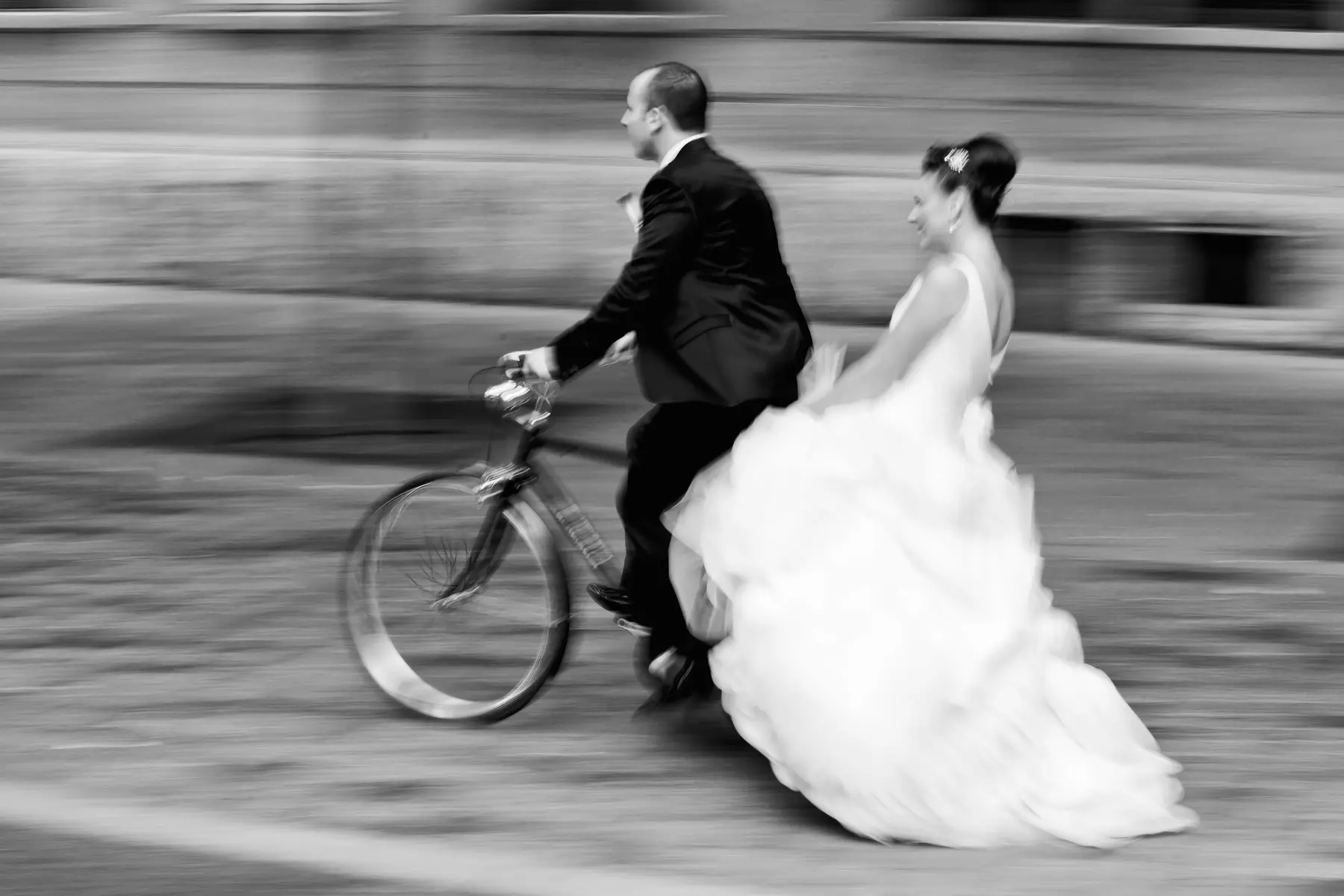 A black and white panning shot shows a groom cycling a bicycle with his bride alongside, her white wedding dress dramatically billowing as they move quickly past a blurred background. Both are smiling slightly.
