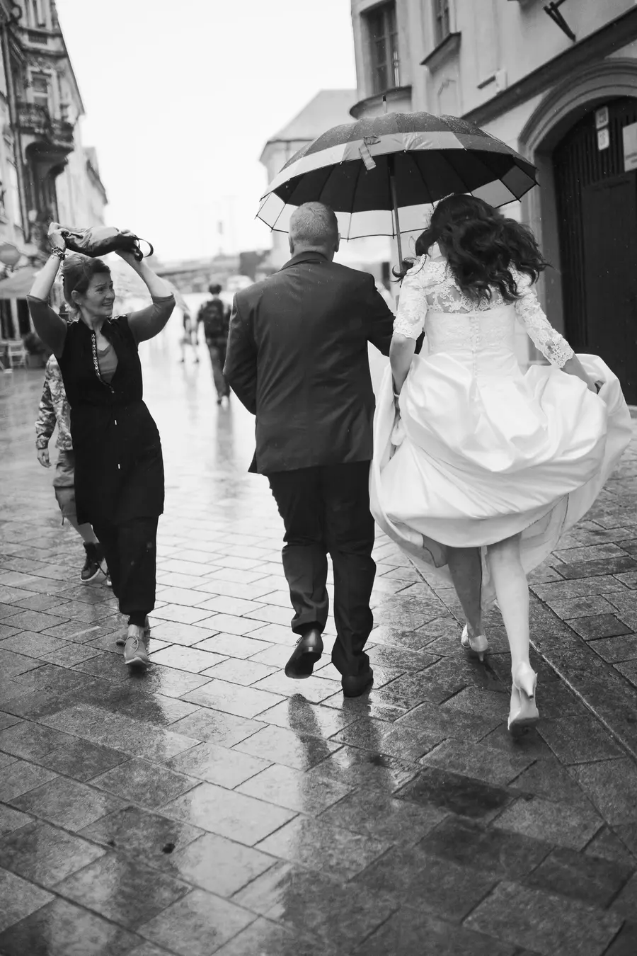 A bride and groom walk under an umbrella on a wet, cobblestone street in the rain. The bride's white wedding dress billows out behind her. A woman walks to their left, holding a bag over her head to shield herself from the rain.