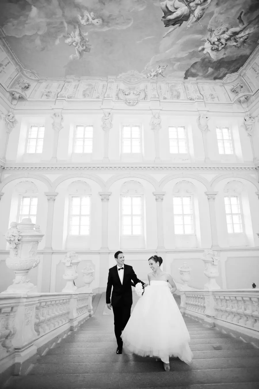 A black and white photo shows a smiling bride and groom descending a grand, ornate white staircase in a richly decorated hall with high windows and a frescoed ceiling.