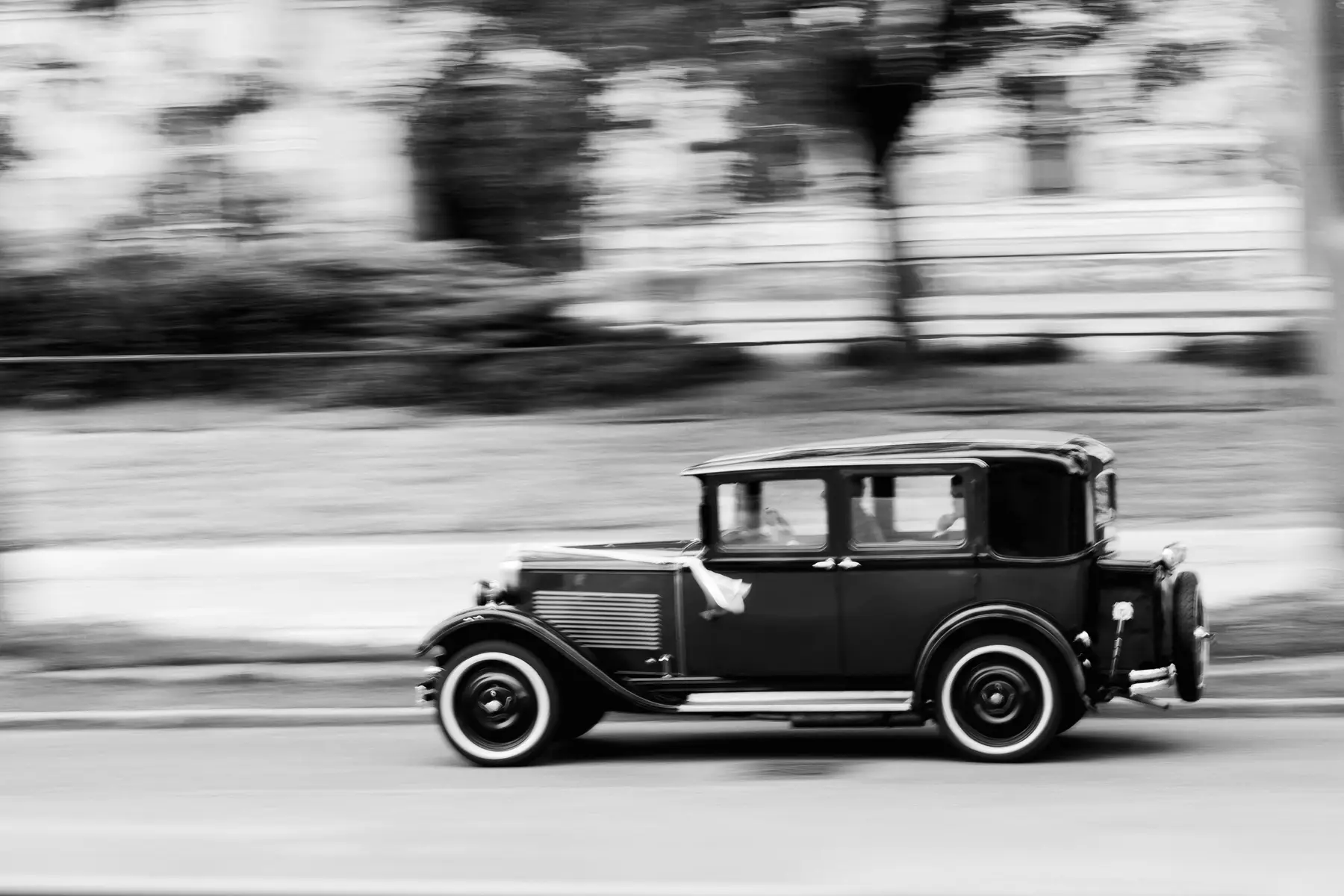 A black and white photo shows a dark vintage sedan with prominent white sidewall tires, driving from left to right. The car is in sharp focus, while the background of trees and grass is blurred horizontally