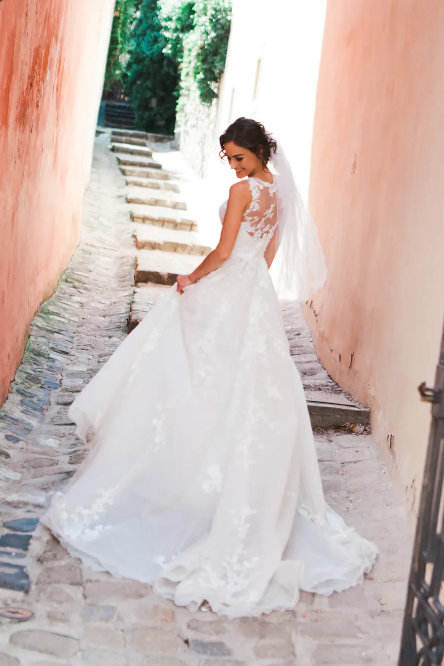 A smiling bride in a white lace wedding dress and veil walks down narrow stone steps in an alley with peach-colored walls, looking back over her shoulder.