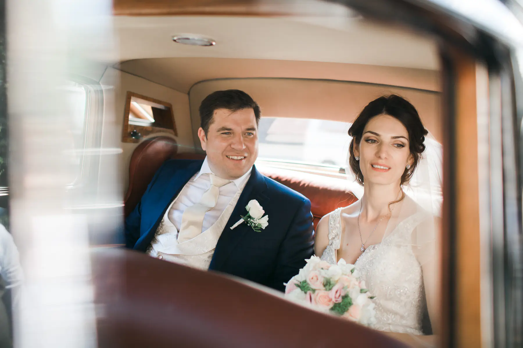 A happy bride and groom are seated in the luxurious back interior of a car. The groom, in a navy suit and cream vest, smiles brightly towards the left, while the bride, in a white