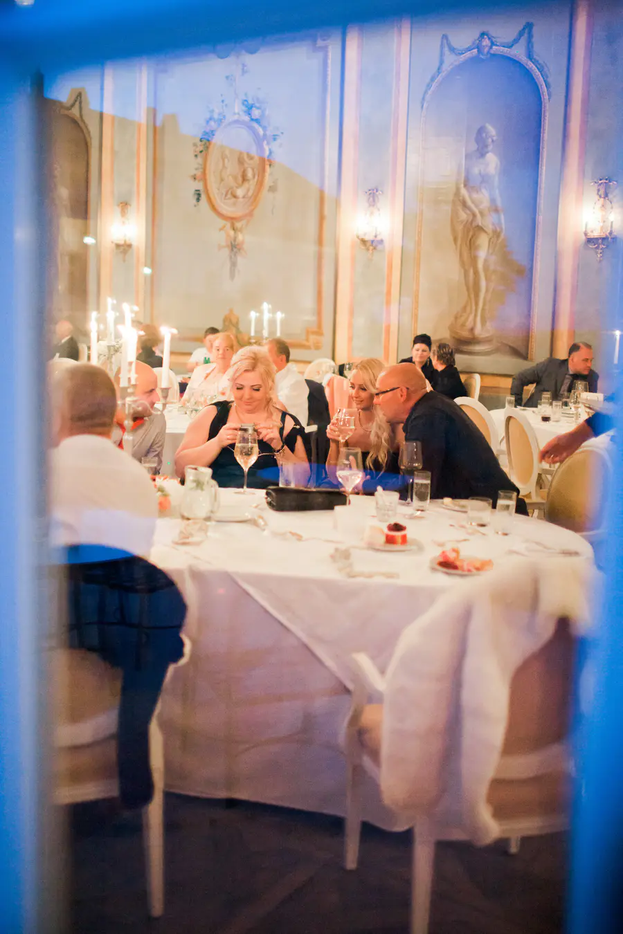 View through a window into an elegant dinner party. Guests are seated at white-clothed tables lit by candles. Classical murals and wall sconces decorate the room's pale walls.
