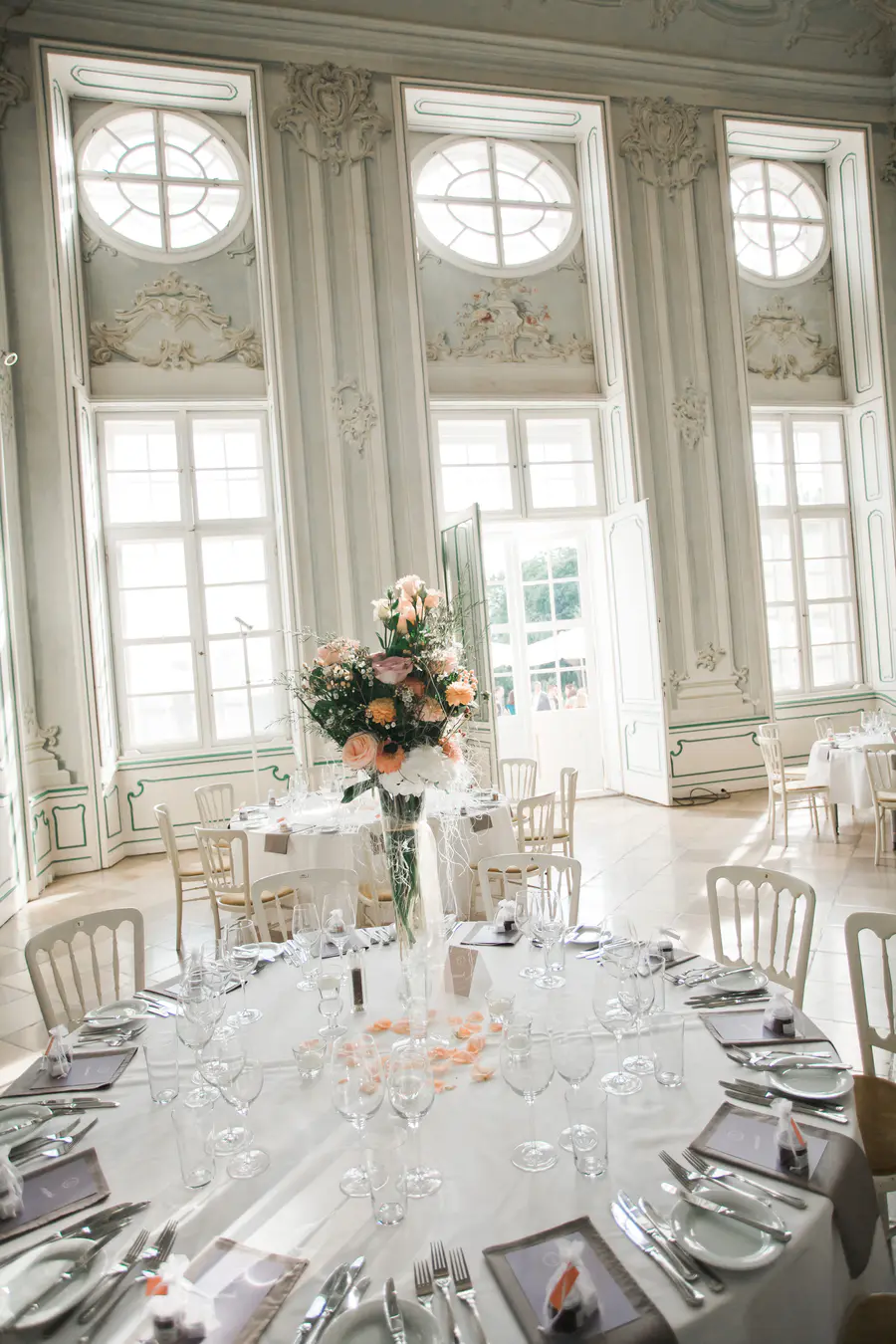 A bright, elegant reception room with multiple tables set with white linens and chairs. A large centerpiece of peach and pink flowers adorns the foreground table. The room features tall windows and ornate pale-colored walls.