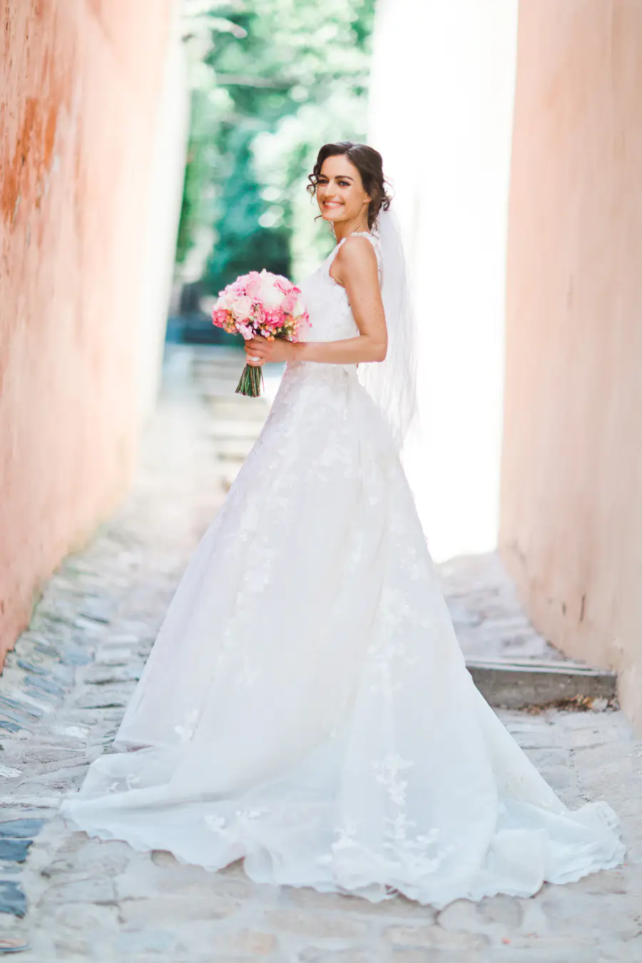 A smiling bride in a white wedding dress stands in a cobblestone alley, holding a bouquet of pink and white flowers.