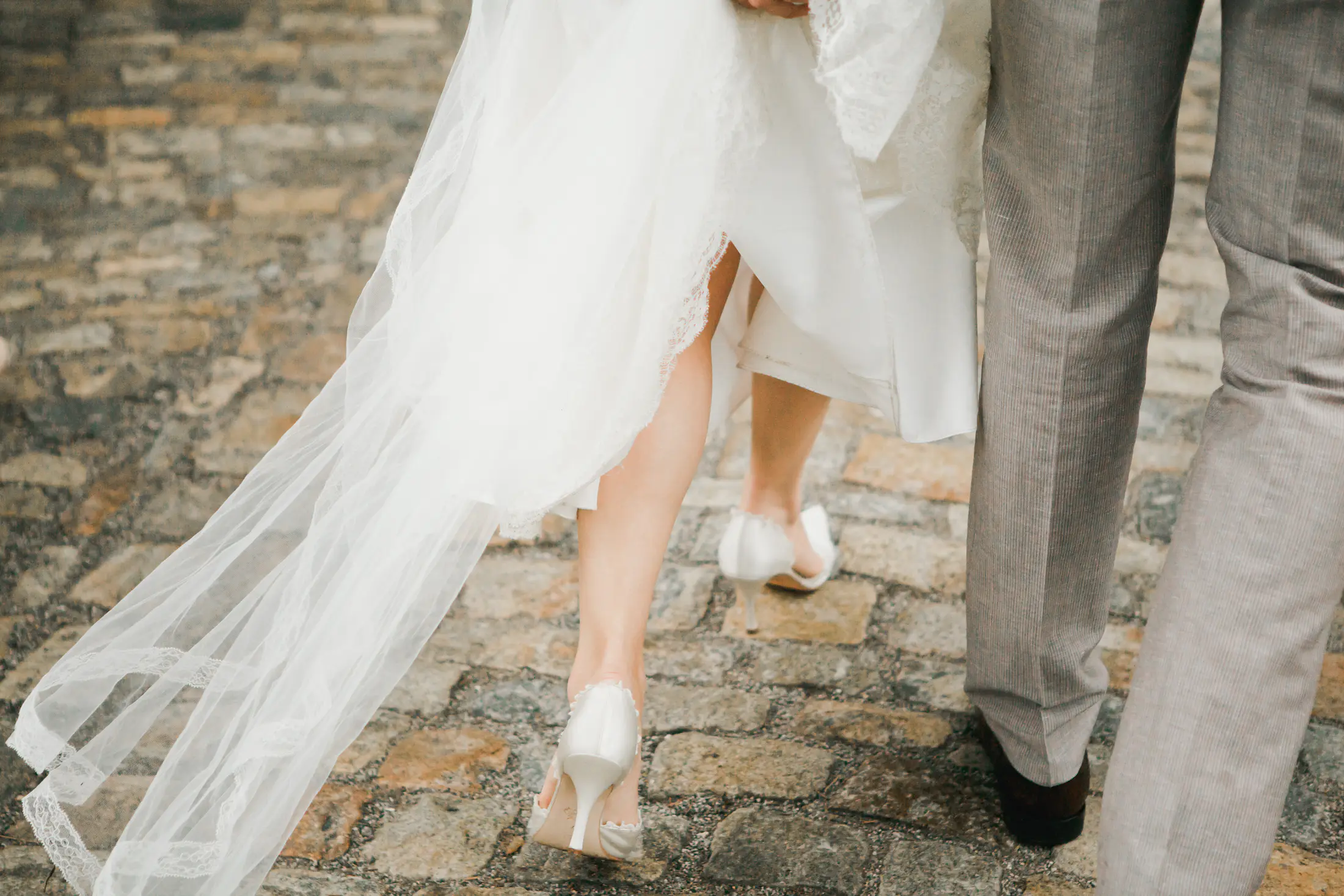 A bride's white wedding dress and veil trail behind her as she walks on a cobblestone path next to a man in gray slacks.