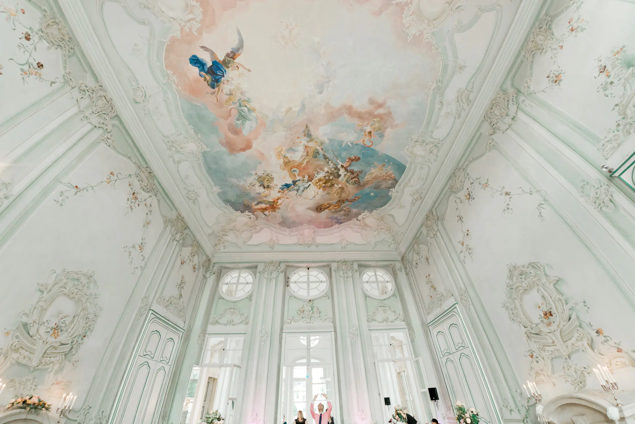 A wide-angle shot shows the ornate interior of a room with a heavily decorated ceiling. The ceiling is painted with a fresco depicting angels and cherubs in pastel colors. The walls are white with light green trim and decorated with delicate floral paintings and intricate white stucco moldings. Large windows with arched tops line the walls, and several candelabras are visible.