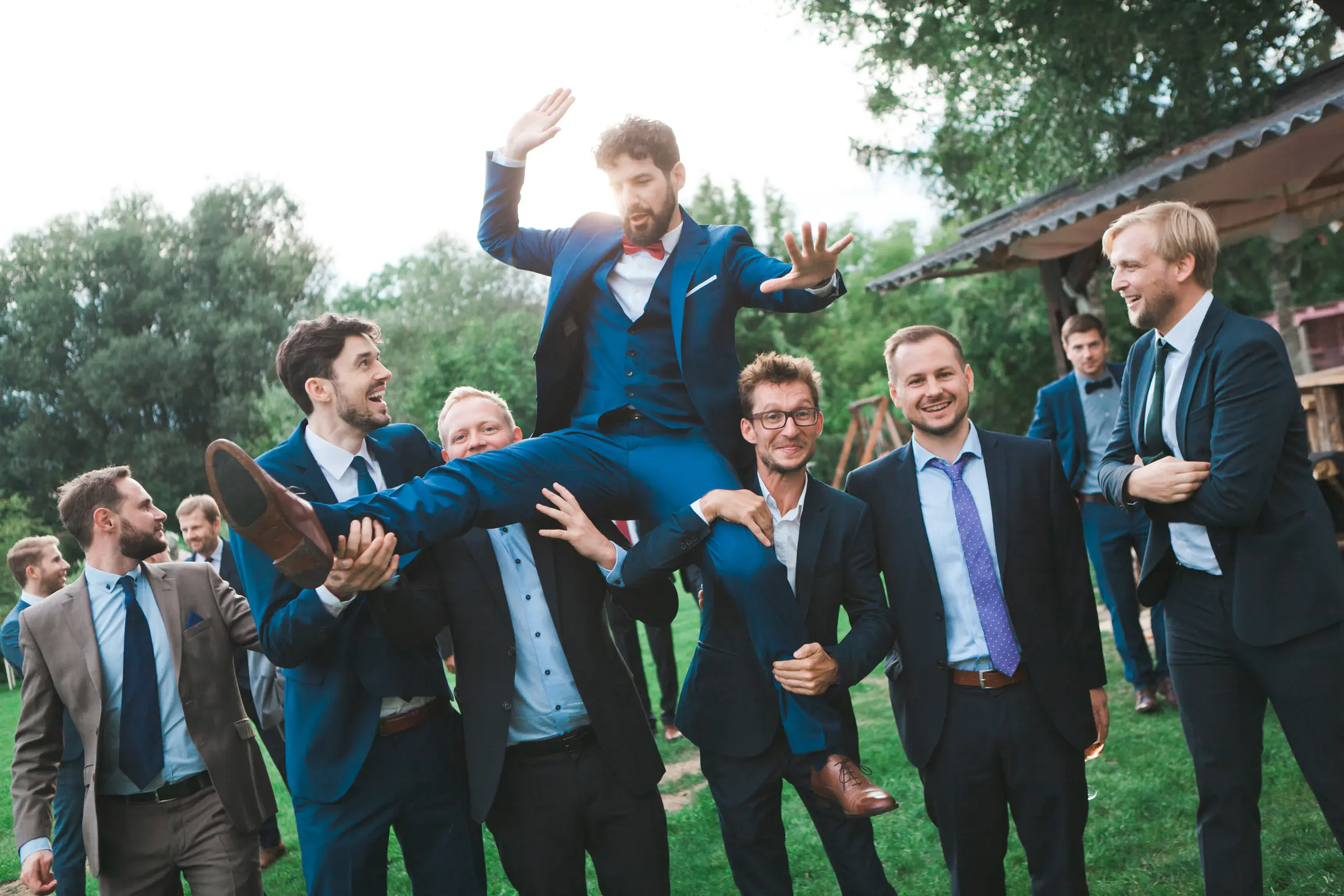 A group of men in suits are celebrating outside. One man, wearing a blue suit and red bowtie, is being carried on the shoulders of two other men.