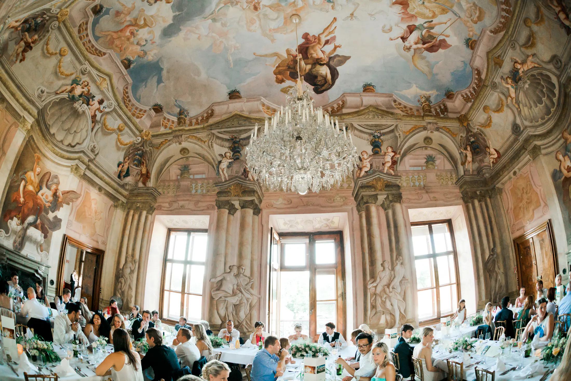 An ornate ceiling mural and a crystal chandelier hang above a wedding reception hall filled with guests seated at long tables.