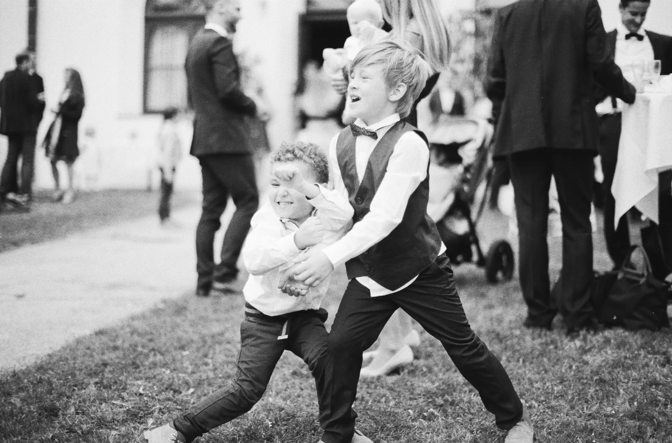 Analoque black and white photo shows two young boys in formal wear playing on grass. One boy with blonde hair is wearing a white shirt, a dark vest, and a bow tie. He is laughing with his mouth open. The other boy, who is smaller with curly hair, is in front and to the left of the blonde boy. He is wearing a white shirt and dark pants. The two boys are holding onto each other. In the background, there are several blurred figures of adults in formal attire.Analoque wedding photography Vienna