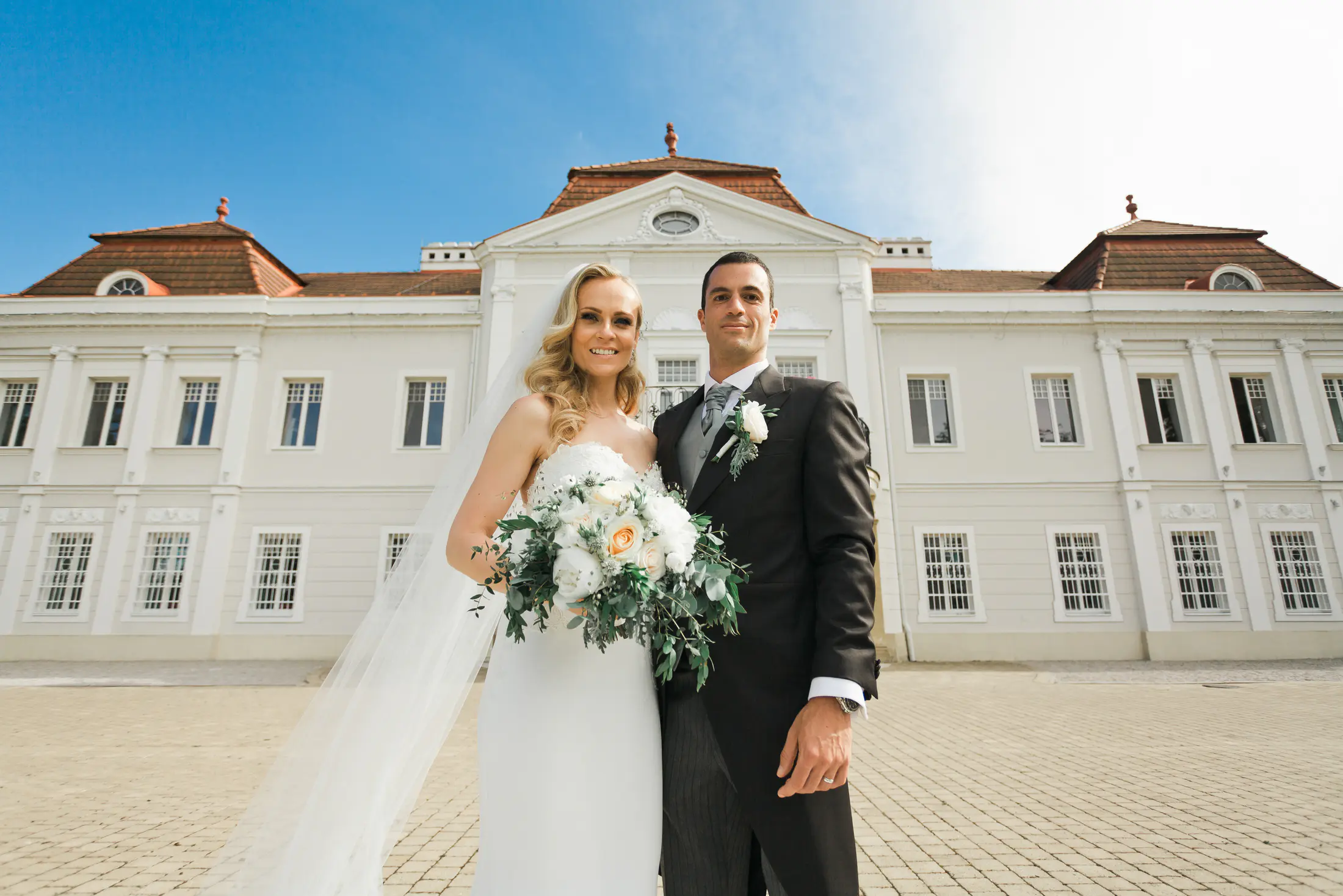 A bride and groom stand in front of a large, white building with a tiled roof. The bride, on the left, wears a white wedding dress and veil and holds a bouquet of white and peach roses. The groom, on the right, wears a black tuxedo with a grey vest and tie. They both smile at the camera. The sky is bright blue.