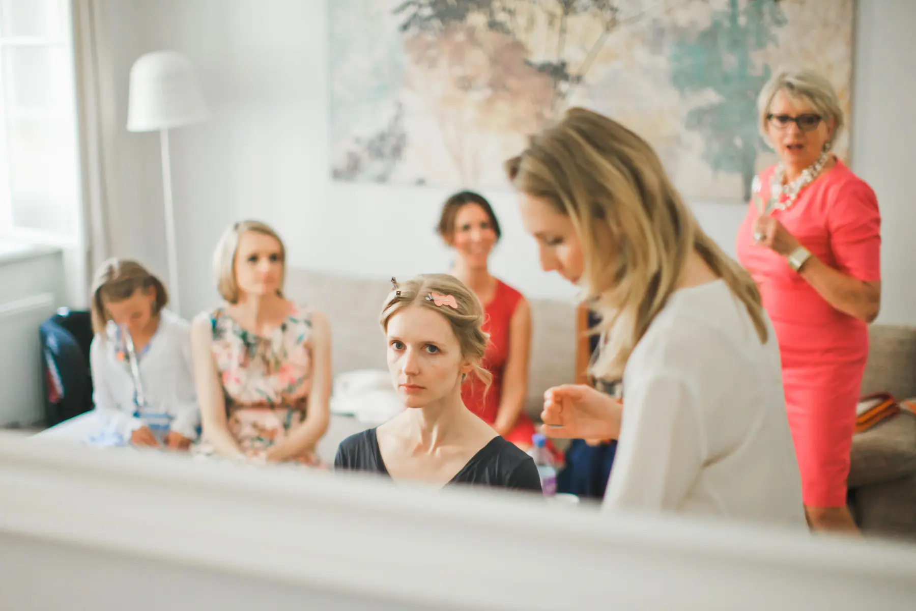 A group of women are gathered in a room, preparing for anwedding. One woman in the foreground is looking directly at the camera, with her hair styled with clips. To her left, another woman in a floral dress is seated, while a third woman in a white robe is visible further back. In the background, a woman in a bright pink dress is speaking to someone out of frame.