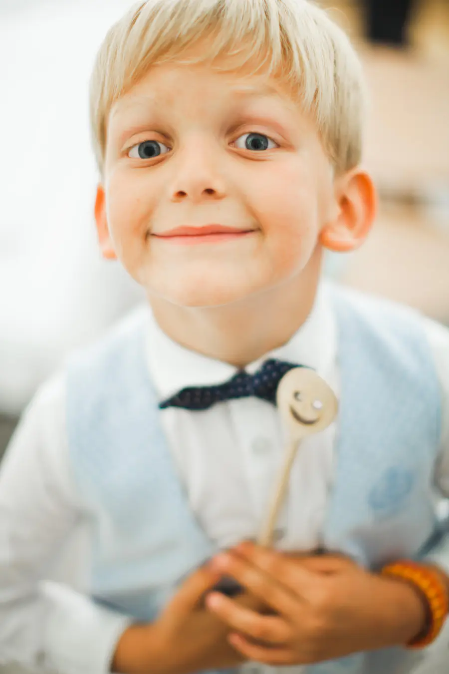 A young boy with blonde hair smiles at the camera while holding a wooden spoon with a smiley face. He is wearing a white shirt, a light blue vest, and a dark blue polka dot bow tie.