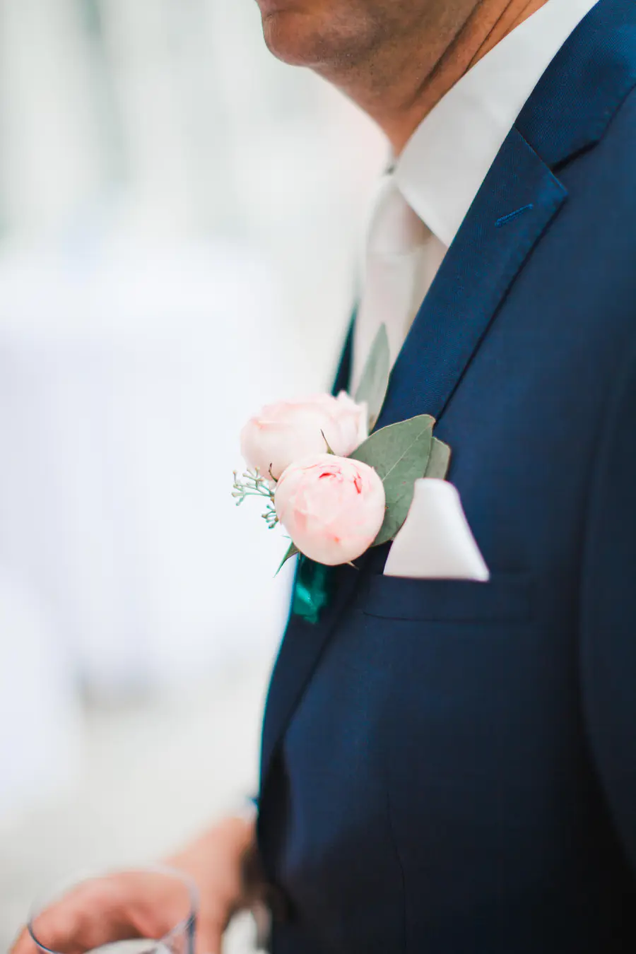A close-up of a person wearing a dark blue suit jacket, white shirt, and light tie. A boutonniere with two light pink roses and green leaves adorns the lapel, and