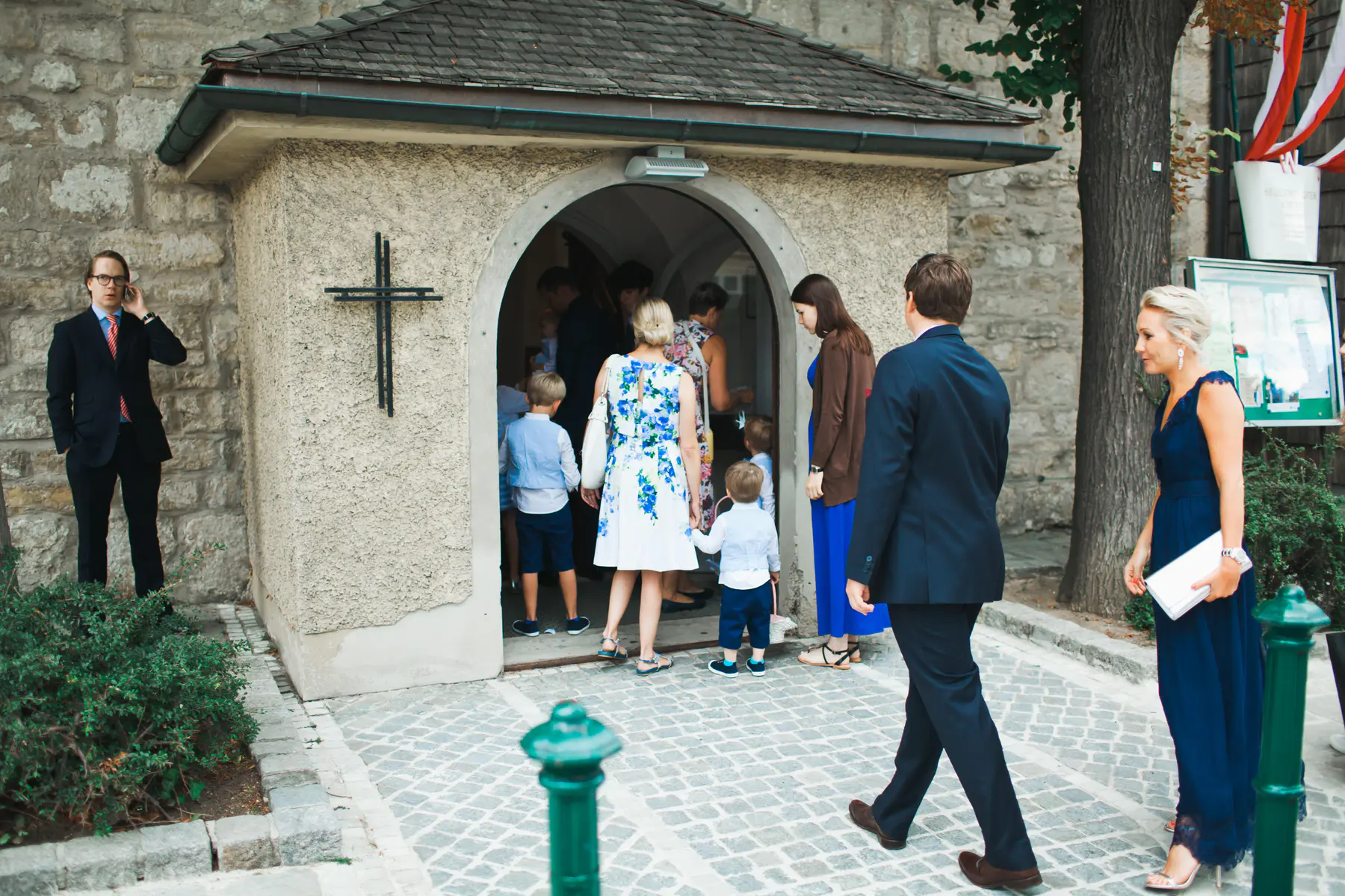 A group of people in formal attire stand outside a stone building with an arched entrance. A man in a dark suit on the left talks on a phone, while several others, including a woman in