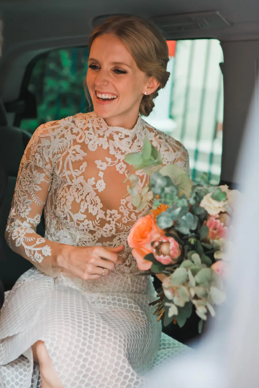 A joyful bride in a white, long-sleeved lace wedding gown is seated in a car, laughing while holding a colorful floral bouquet.