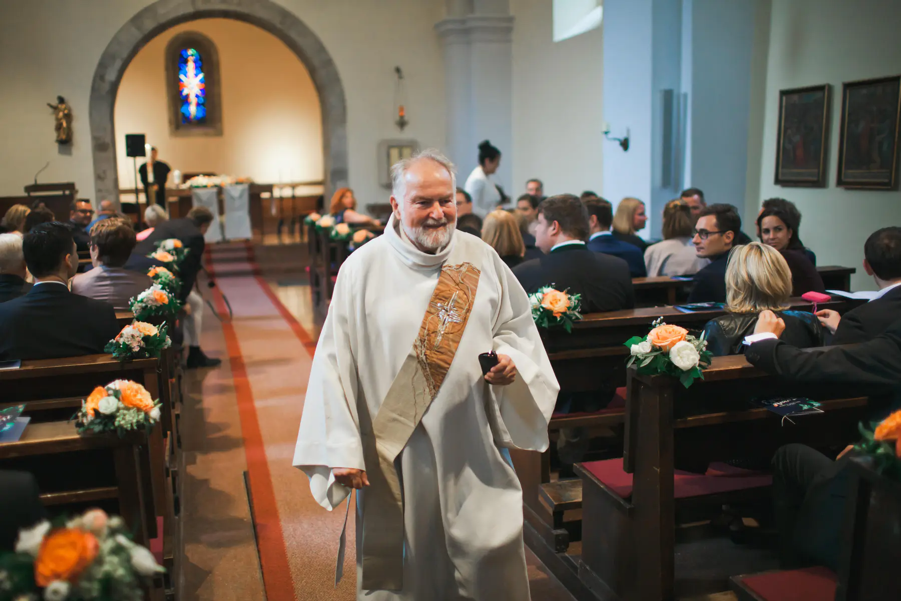A smiling man in a white clerical robe with an embroidered stole walks down the central aisle of a church. Seated attendees line the pews on both sides, which are decorated with small bouquets of orange