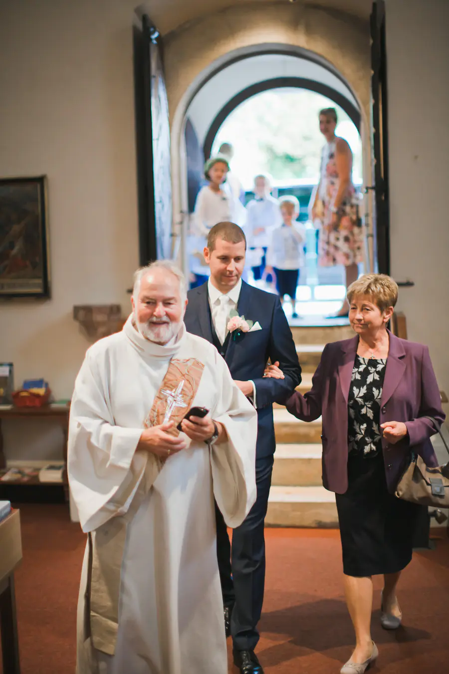 A smiling priest in white robes walks next to a groom in a suit and an older woman in a purple jacket, holding his arm. More guests are visible in the bright background through an arched church doorway