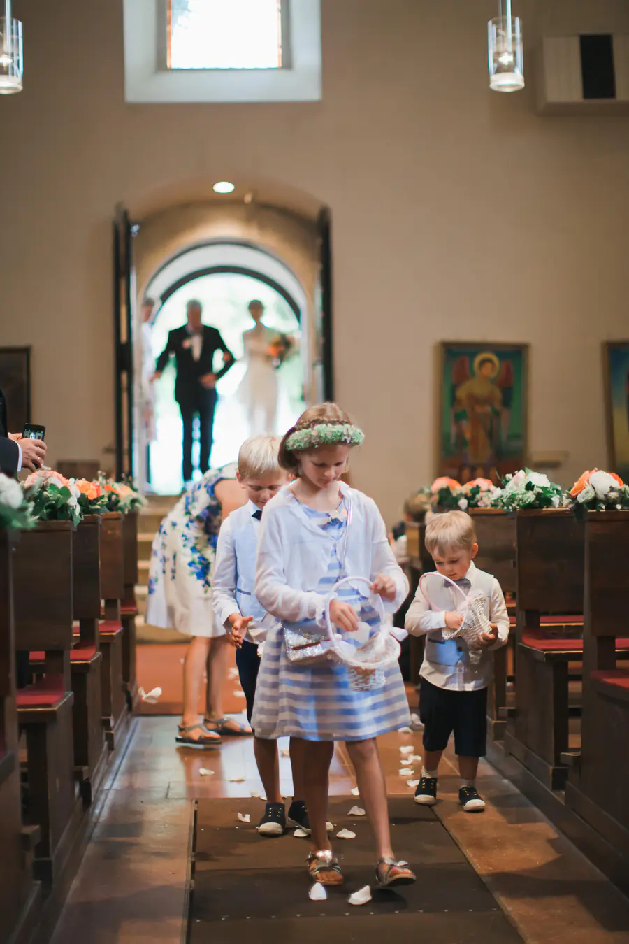 A young flower girl in a striped dress and flower crown, followed by two page boys, scatters white petals down a church aisle. In the blurred background, a bride in a white gown and a