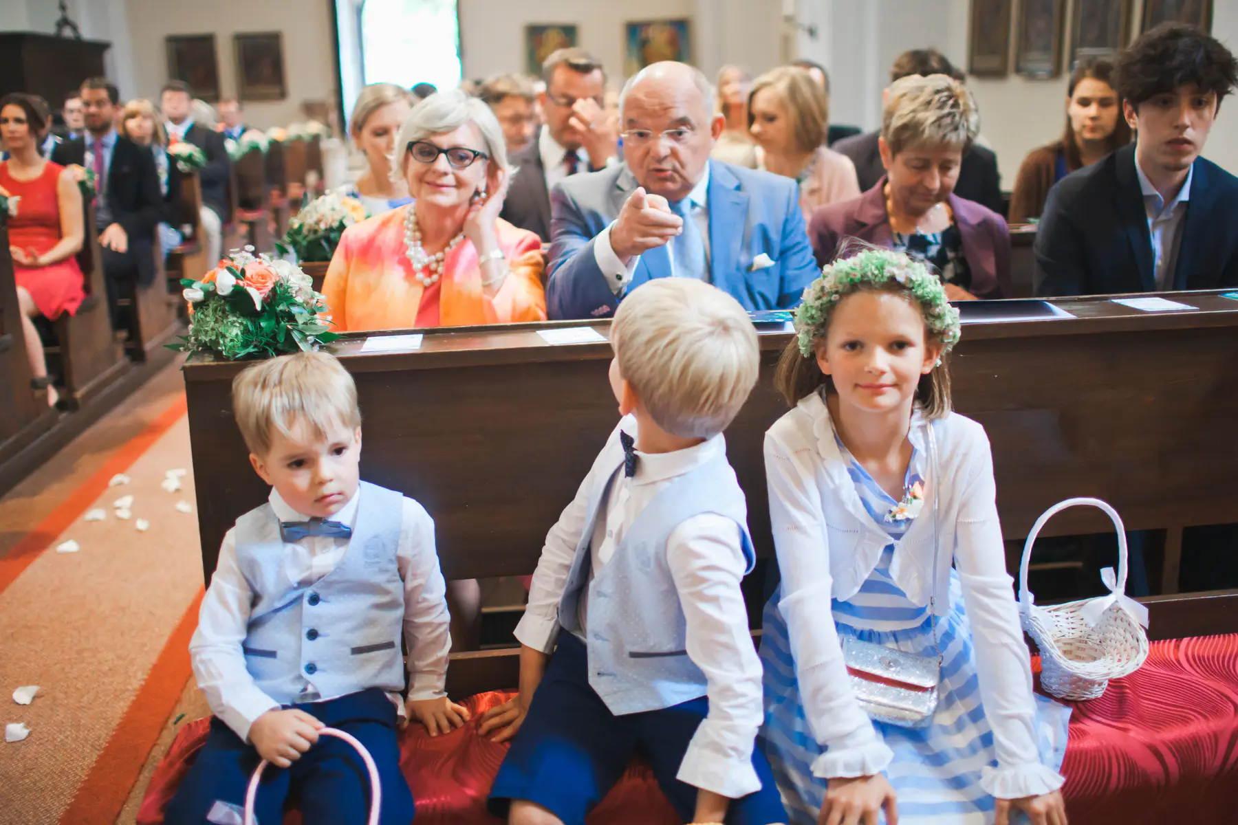 Three children, likely a flower girl in a floral crown and two ring bearers in vests, sit in the front pew of a church during what appears to be a wedding. Adults are seated in the pews behind them.
