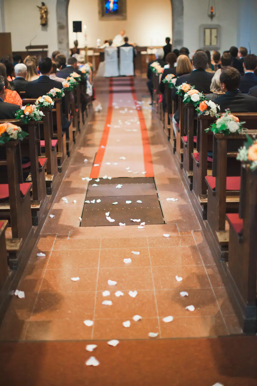 A view from the back of a church aisle during a wedding ceremony. Guests are seated in wooden pews on both sides, which are decorated with small bouquets of white and orange flowers.