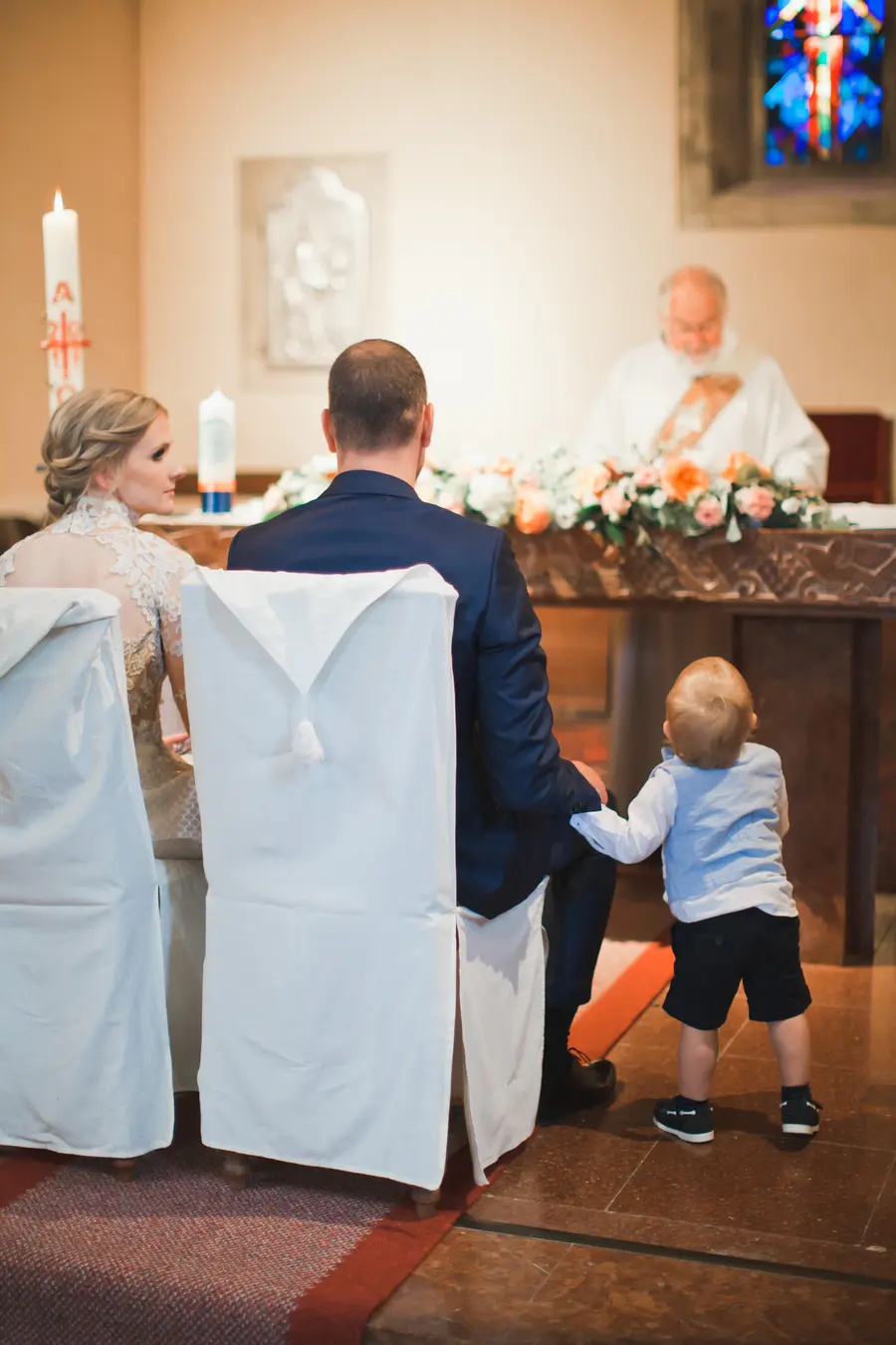 A wedding ceremony in a church, viewed from behind the bridal couple and a child. The groom, in a dark suit, sits with a toddler holding onto his leg. The bride,