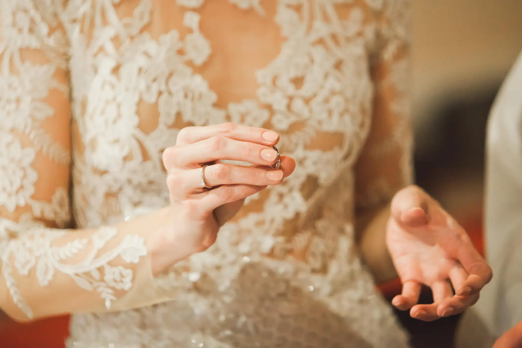 A bride wearing a white lace wedding dress holds a plain silver ring between the thumb and index finger of her left hand. A diamond engagement ring is on her left ring finger, and her right hand is open, palm up.