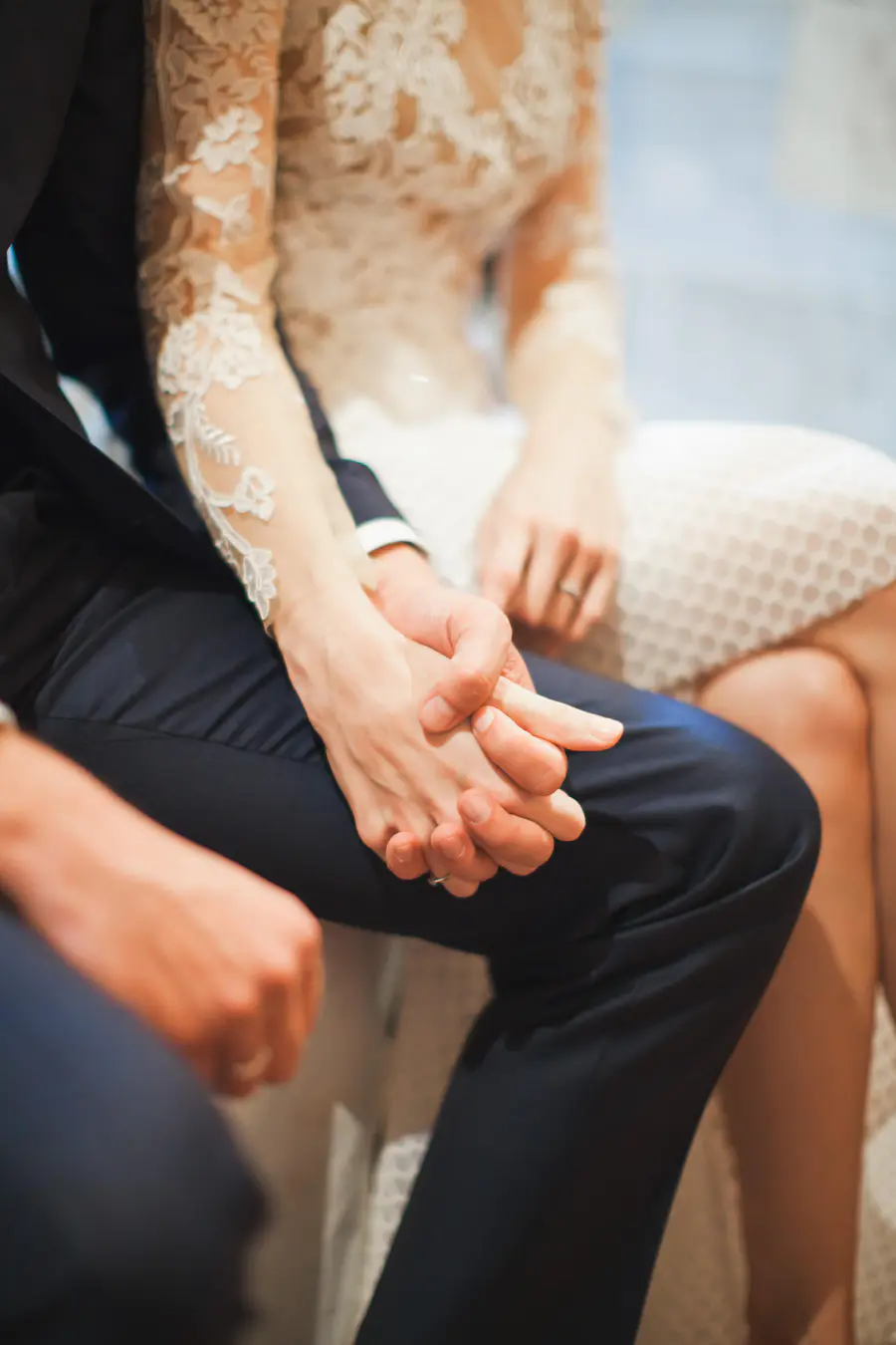 A close-up shot of a bride and groom holding hands. The bride wears a white lace dress, and the groom wears a dark suit. Both wear wedding rings.