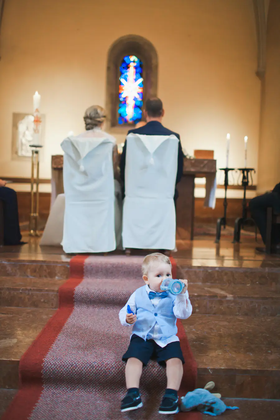 A toddler in a blue vest and bow tie sits on a red carpeted church step, drinking from a sippy cup, with a small blue plush toy beside him. In the background, two adults
