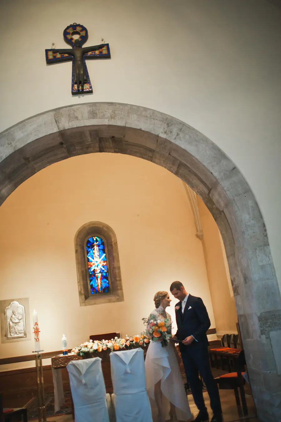 A bride in a white high-low wedding dress, holding a peach and orange bouquet, stands facing her groom in a dark suit and bow tie. They gaze at each other intimately inside a church,