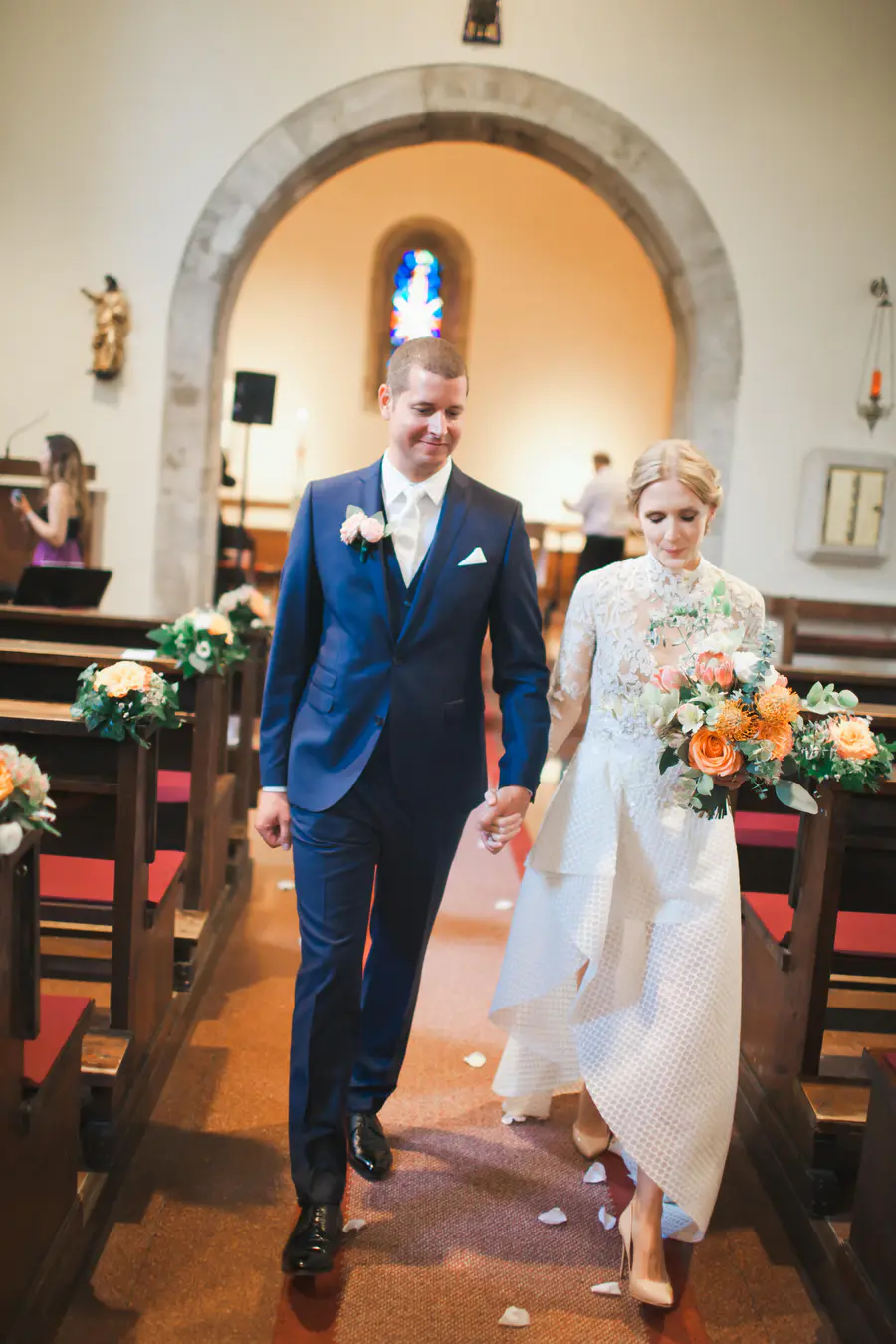 A newlywed couple walks hand-in-hand down a church aisle scattered with white rose petals. The groom, in a dark blue suit, smiles as he looks down. The bride