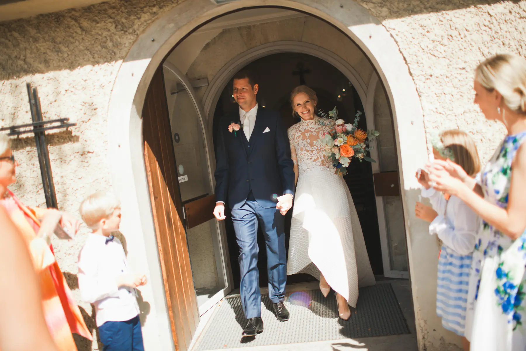 A beaming bride in a white lace dress holding a colorful bouquet and a groom in a navy suit exit an arched stone church door, holding hands, as sunlight streams down and guests watch from the sides.
