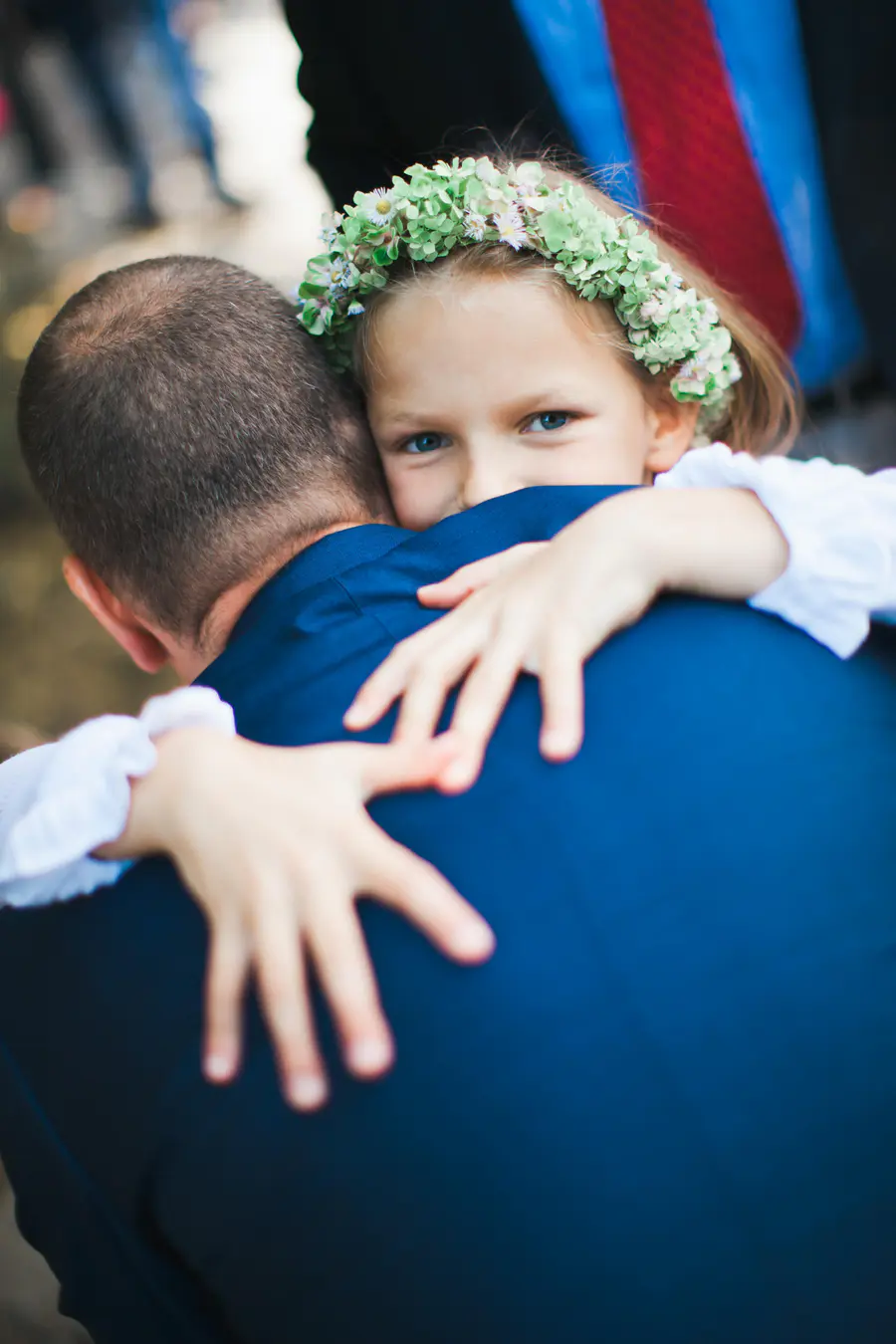 A child wearing a green flower crown hugs an adult from behind, looking directly at the viewer over their shoulder. The adult is seen from the back, wearing a dark blue suit jacket.