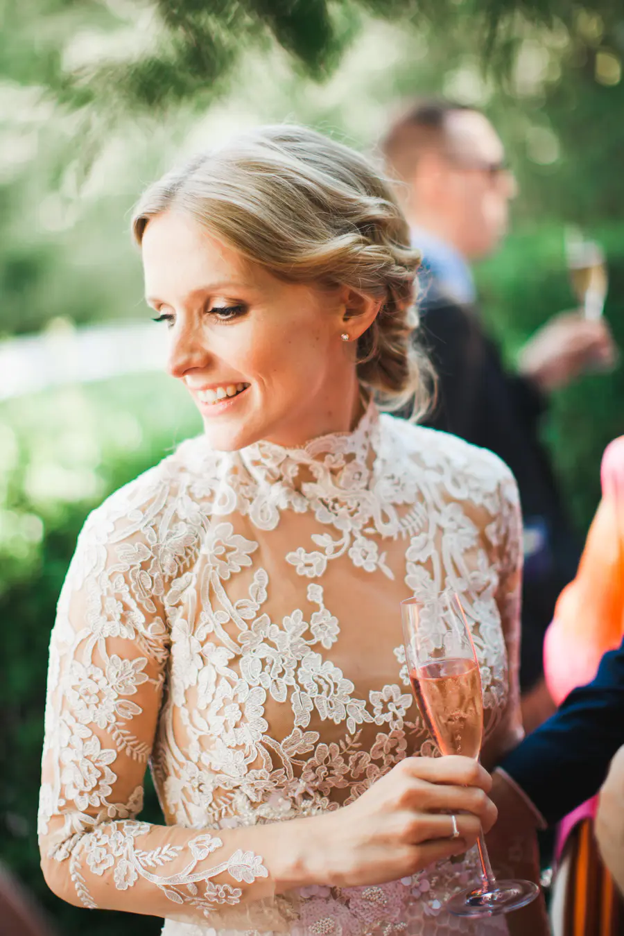 A smiling bride in a white lace dress holds a glass of champagne.