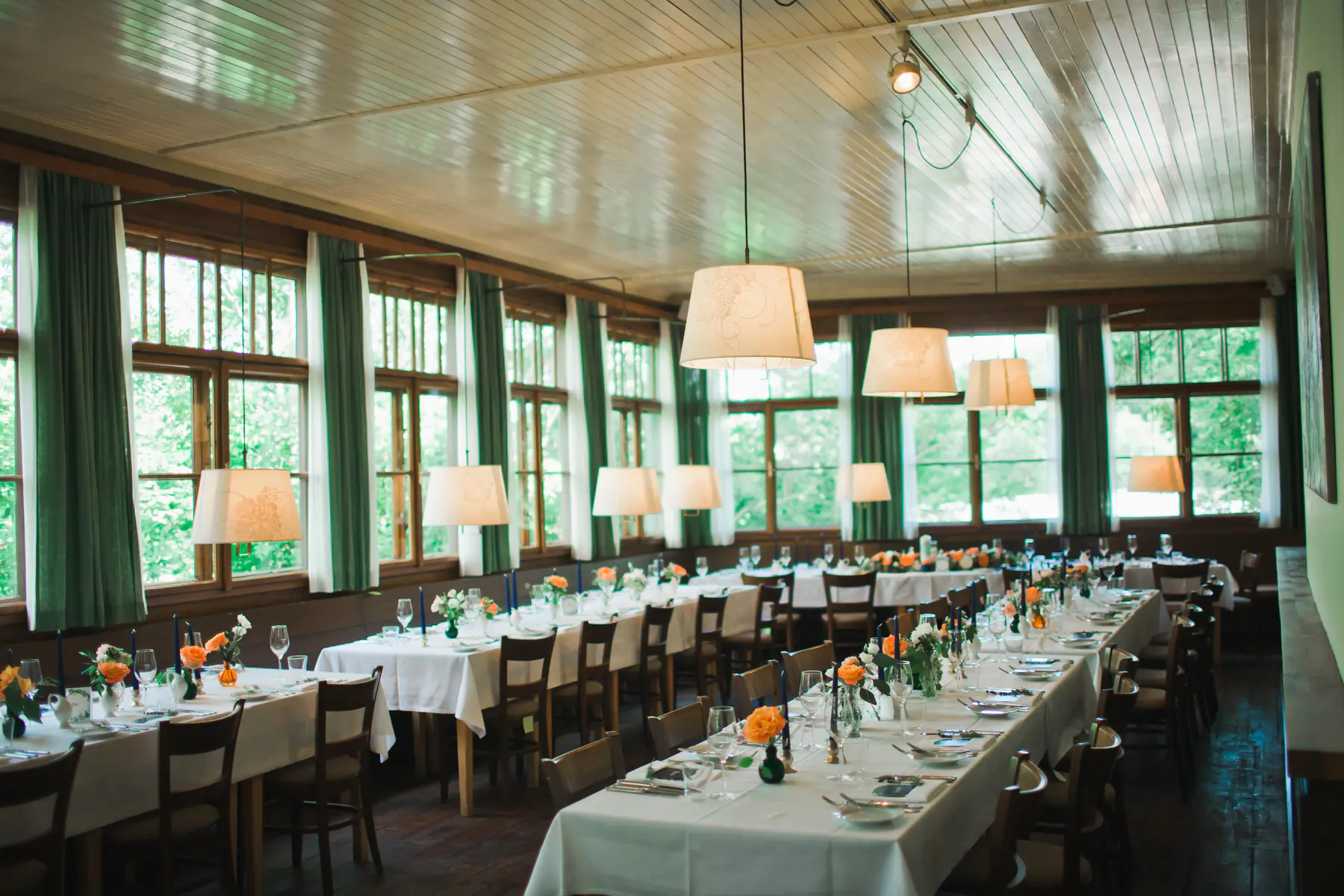 Rustic wedding elegance in the historic halls of Pfarrwirt. A long dining room with multiple tables set for a formal event, with white tablecloths, floral centerpieces, and hanging lamps.