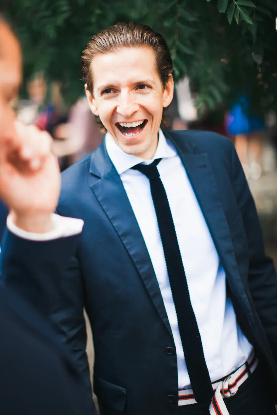 A man with slicked-back dark hair in a navy suit, white shirt, and black tie is laughing joyfully, looking towards a blurred figure in the foreground. Blurred green foliage is visible in