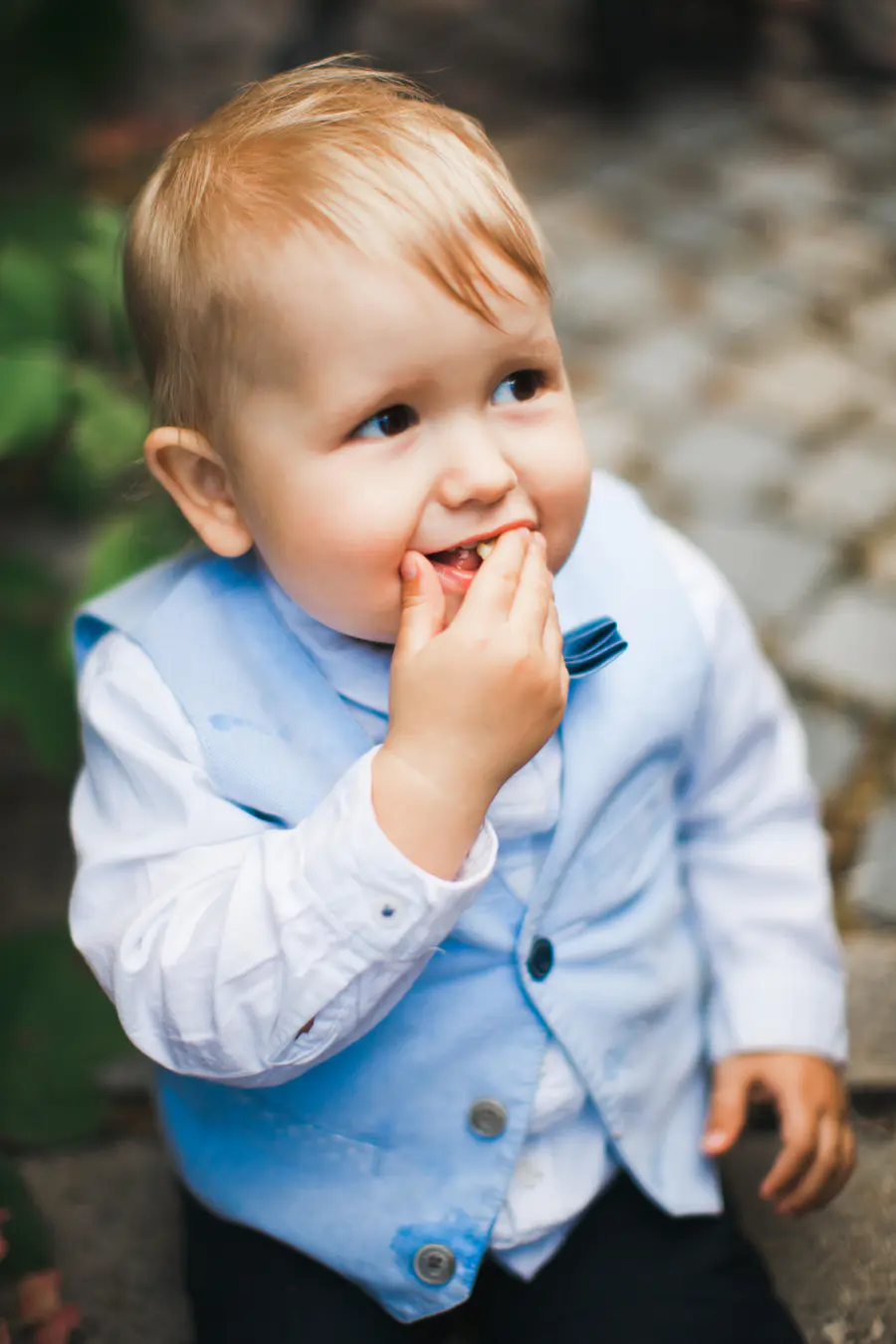 A toddler with blonde hair wears a white shirt and a light blue vest with a matching bow tie, and puts his fingers in his mouth.