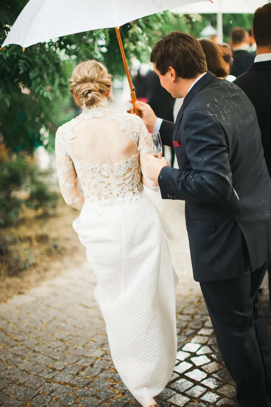 A bride and groom walk together under a white umbrella. The bride wears a white lace wedding dress and her hair is styled in an updo. The groom is in a dark suit and holds a glass of champagne.