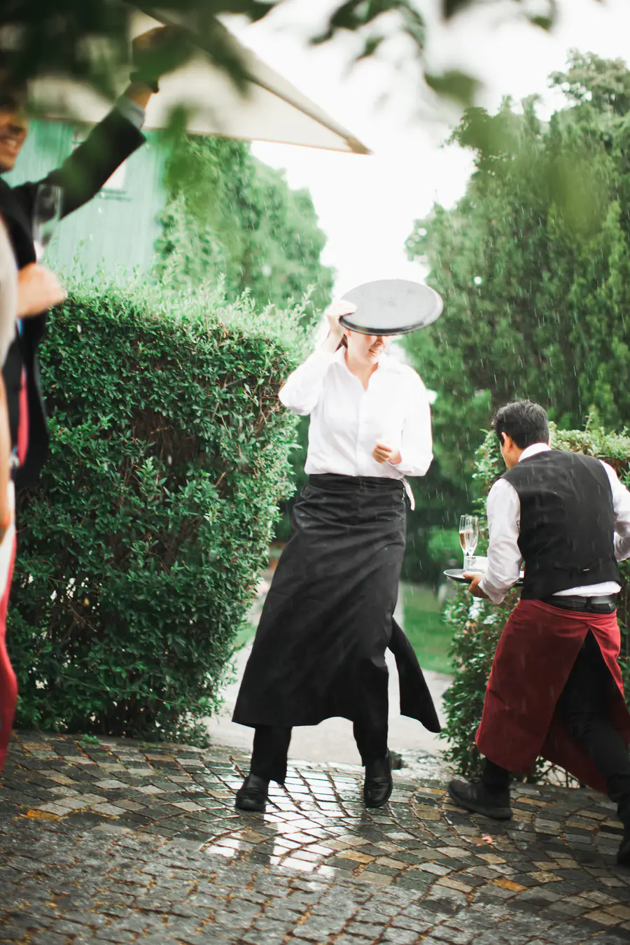 A server in a white shirt and black apron holds a tray over their head as it rains. Another server in a vest and red apron walks by carrying a tray with drinks.