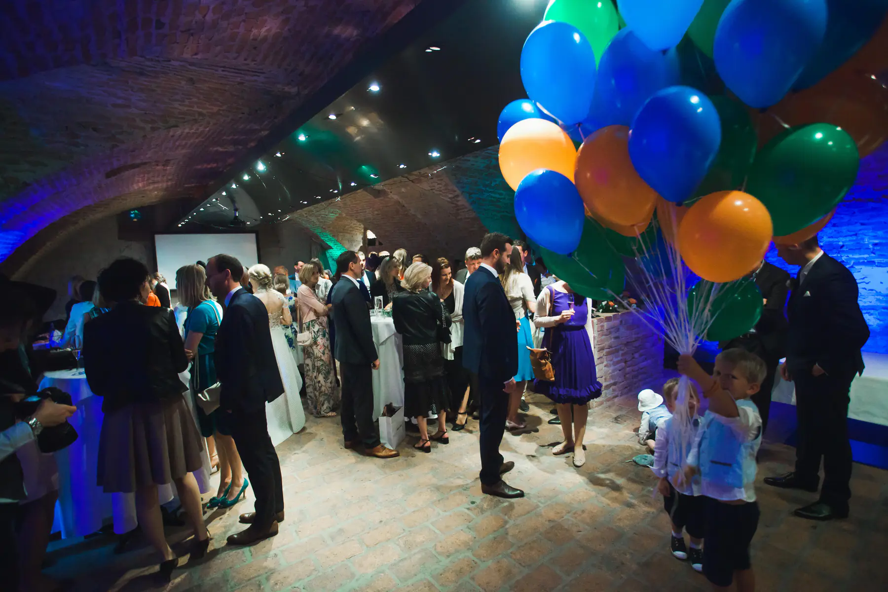 A young boy holds a bunch of blue, orange, and green balloons at a party. Several adults in formal wear are gathered in a room with exposed brick walls and arched ceilings, illuminated by colored lights.