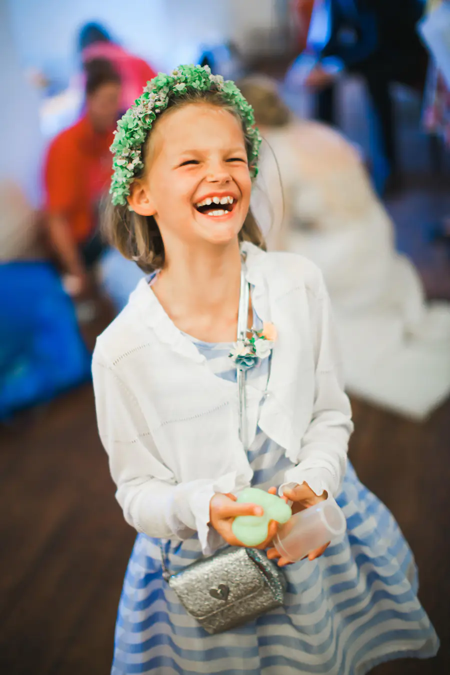A young girl wearing a green flower crown laughs heartily. She is dressed in a white cardigan over a blue and white striped dress, and carries a small silver purse.