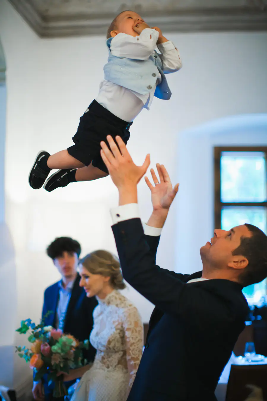 A man in a suit throws a laughing baby boy in the air.Authentic wedding reception at Pfarrwirt Vienna – candid shots