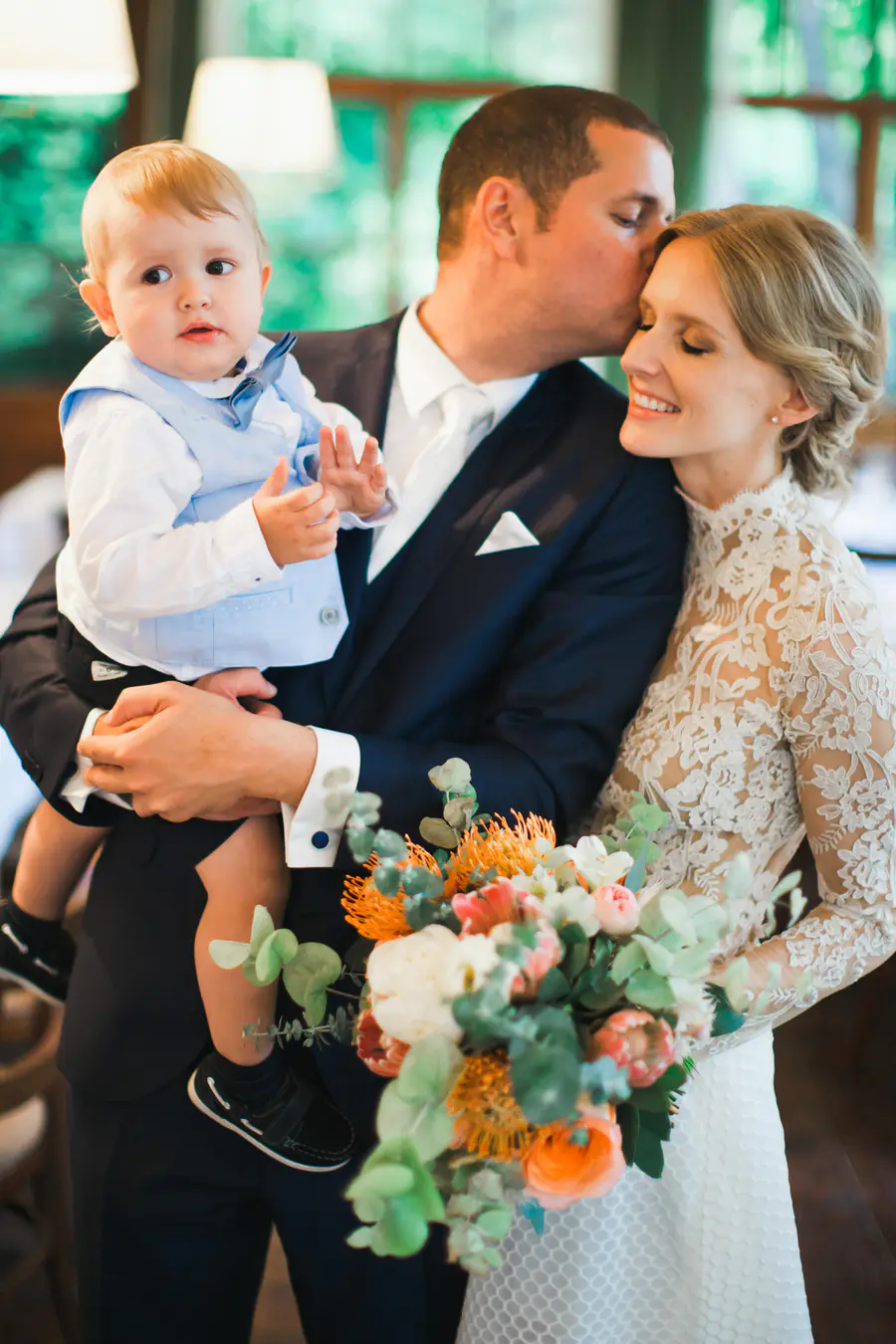 A bride, groom, and baby pose for a photo, with the groom kissing the bride's forehead while holding the baby. The bride smiles and the baby looks away from the camera. The groom is wearing a navy suit, the bride a white lace wedding dress, and the baby a light blue vest and bowtie. A bouquet of flowers is held in front of them.