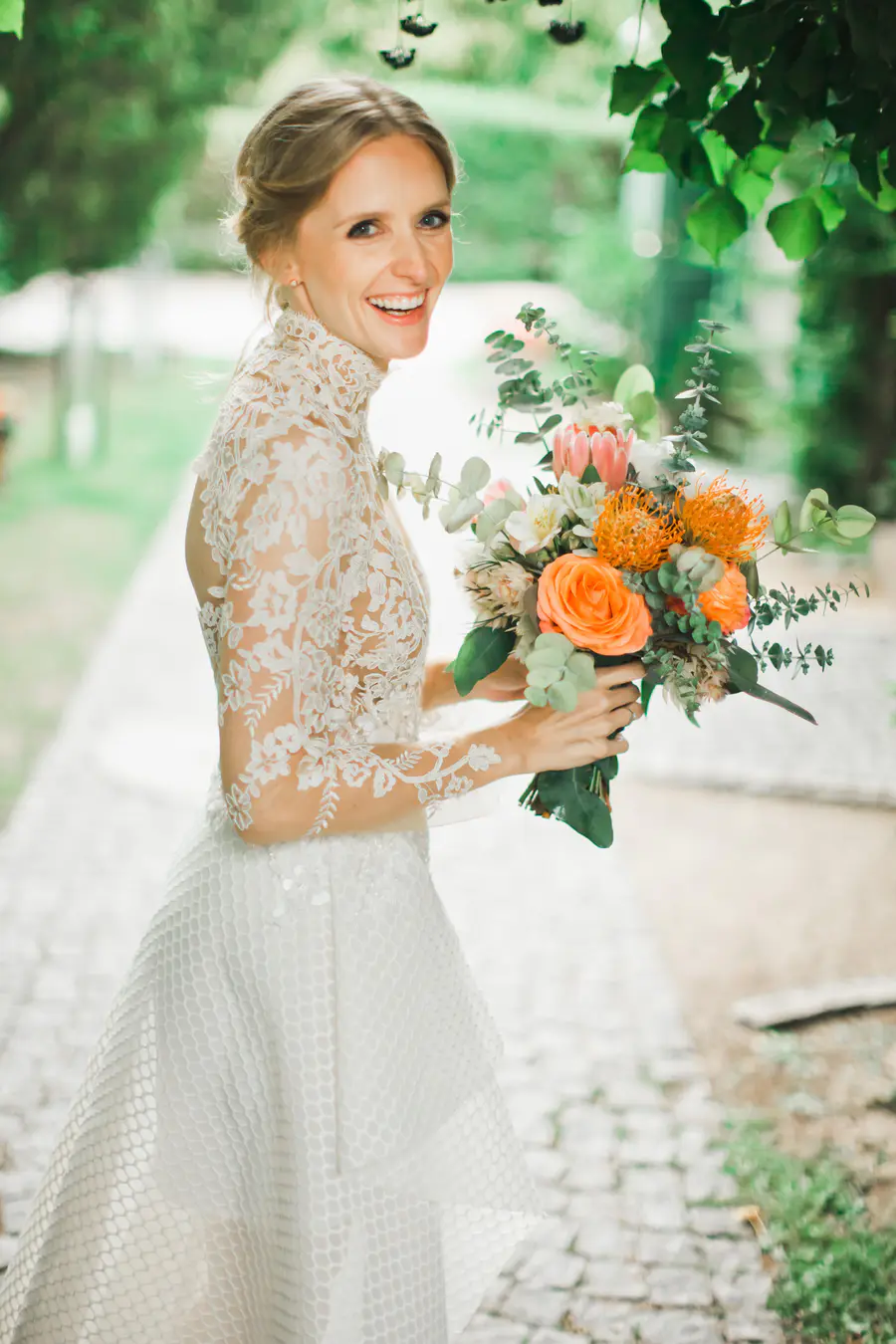 A smiling bride with bright blue eyes and light brown hair styled in an updo, wearing a white lace wedding dress and holding a colorful bouquet.