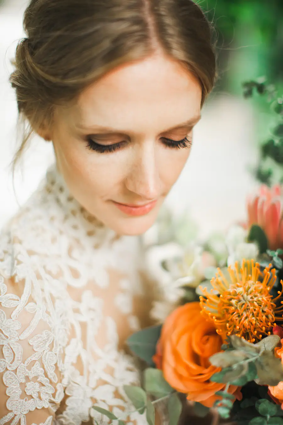 A close-up portrait of a bride in a white lace dress, looking down at an orange and pink bouquet.