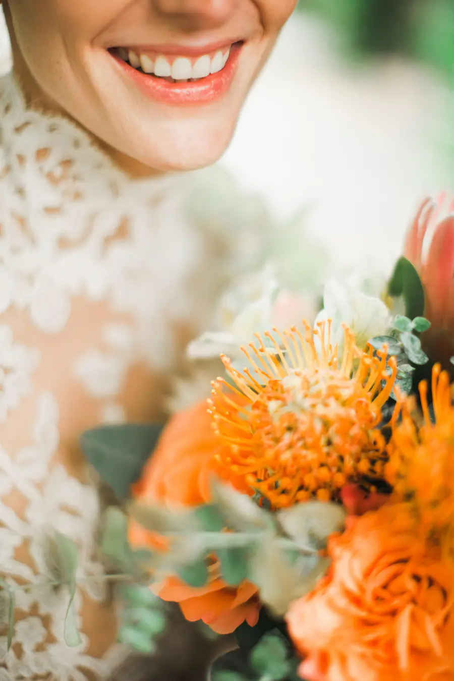 Close-up of a bride smiling, her mouth showing teeth. She is wearing a white lace dress and holding an orange and pink bouquet of flowers.