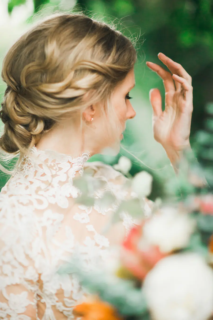 A bride with blonde hair in an updo, wearing a white lace dress, looks to the side with her hand near her face.