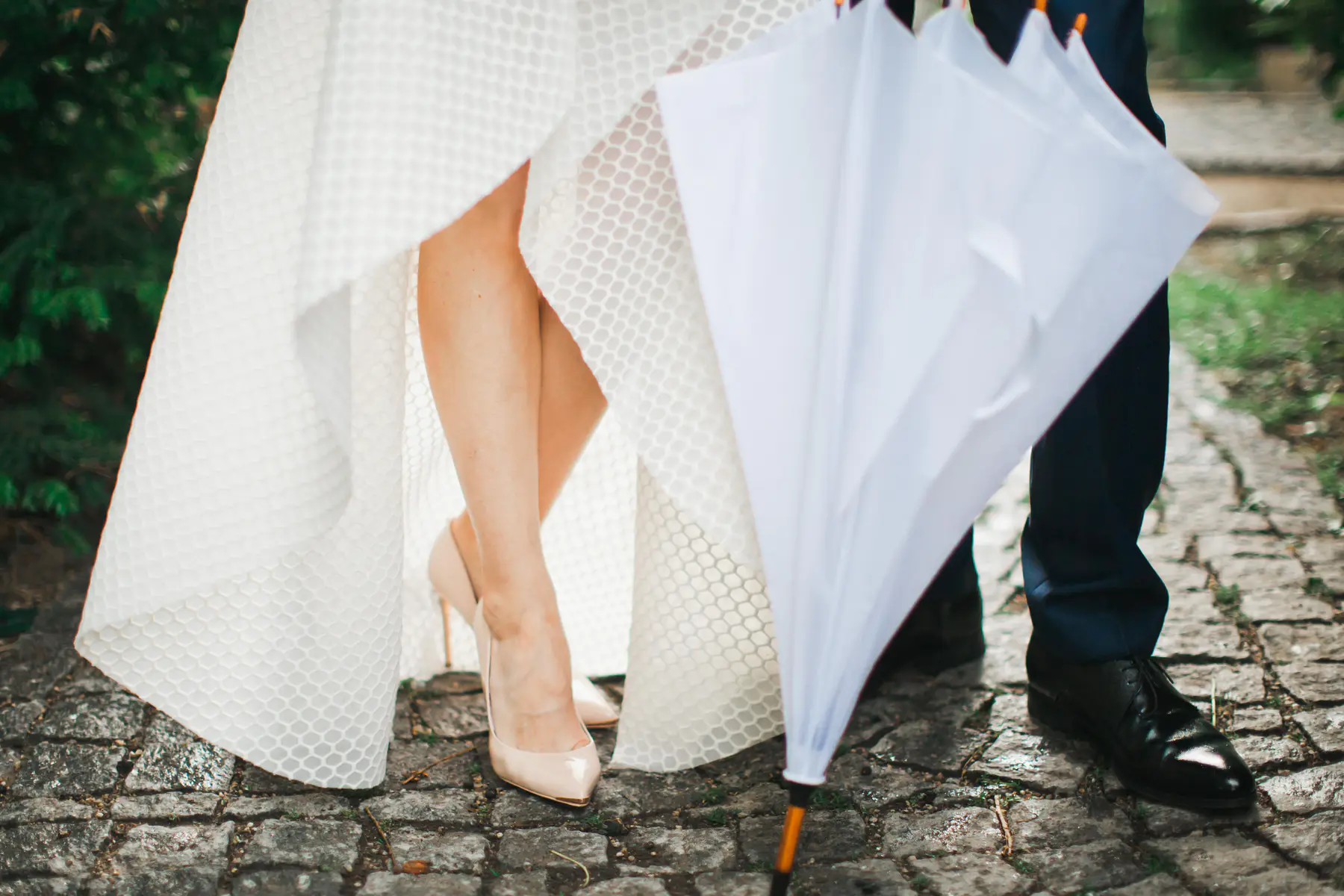 A bride in a white dress and heels stands next to a groom in a suit and black shoes. They are holding a white umbrella and standing on a cobblestone path.