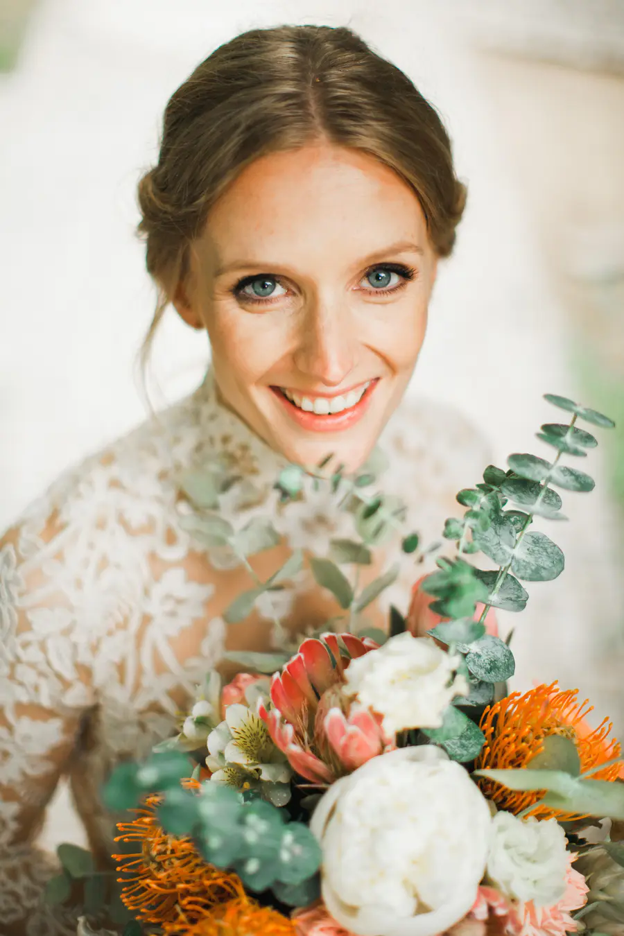 A close-up portrait of a smiling bride with blue eyes and blonde hair pulled back. She is wearing a white lace wedding dress and holding a colorful bouquet of flowers.
