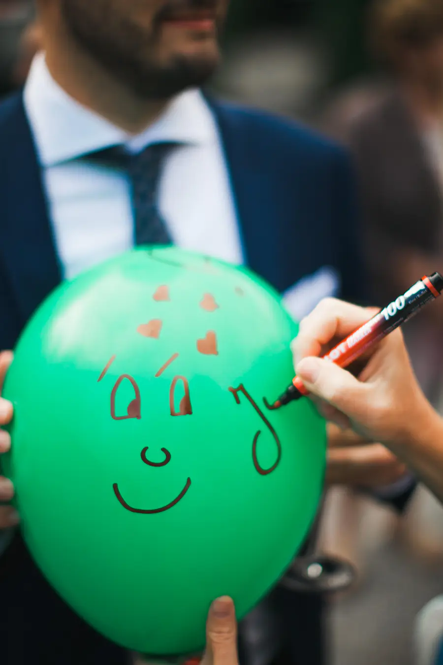 A person's hand is drawing a smiley face with a red marker on a green balloon.